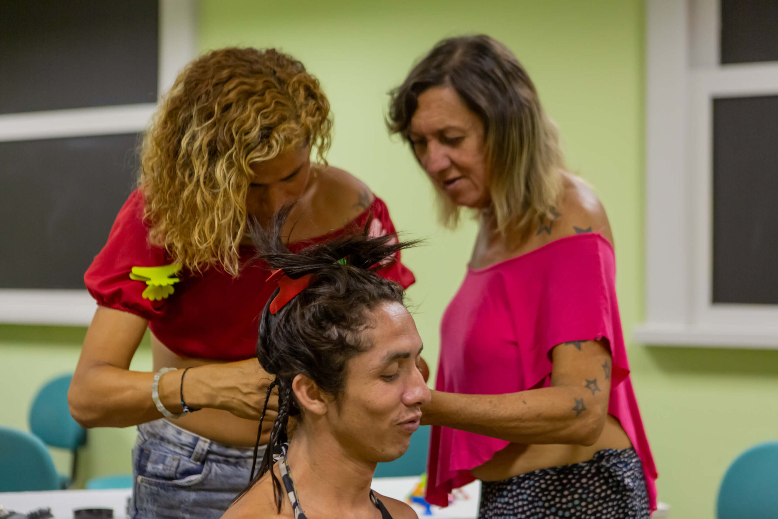 Foto com a presença de três mulheres. Uma delas está sentada, enquanto as outras duas estão trançando seu cabelo.