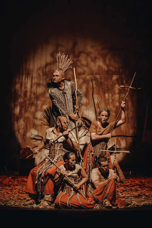 Fotografia de Brunno Martins de uma trecho do espetáculo 1877, da Trapiá Cia Teatral. Nela vemos 5 pessoas, entre homens e mulheres num cenário árido, com vestes cor terra, e detalhes em folhas secas e elementos da natureza, seus rostos estão cobertos de barro e em suas mãos seguram uma cruz de gravetos.