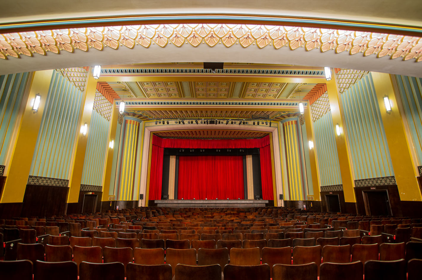 A imagem mostra o interior do Cineteatro São Luiz. Ele tem um estilo arquitetônico clássico. A sala possui cadeiras de madeira dispostas em fileiras, com um corredor central. O teto e as paredes apresentam detalhes ornamentados com padrões geométricos e cores vibrantes, incluindo amarelo, azul e dourado. O palco está ao fundo, decorado com uma grande cortina vermelha fechada, cercada por molduras ornamentais. As luminárias nas paredes complementam o ambiente com uma iluminação suave, destacando os detalhes decorativos.